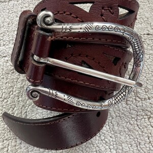 Peut inclure: Une ceinture en cuir marron fonc&eacute; avec une boucle argent&eacute;e d&eacute;corative. La ceinture pr&eacute;sente des formes g&eacute;om&eacute;triques d&eacute;coup&eacute;es et une boucle argent&eacute;e d&eacute;taill&eacute;e avec des volutes. La ceinture est pr&eacute;sent&eacute;e sur une surface textur&eacute;e de couleur claire.