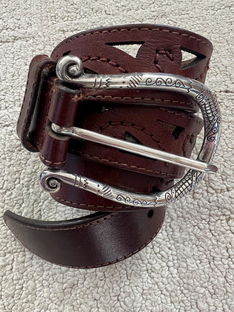 Peut inclure: Une ceinture en cuir marron fonc&eacute; avec une boucle argent&eacute;e d&eacute;corative. La ceinture pr&eacute;sente des formes g&eacute;om&eacute;triques d&eacute;coup&eacute;es et une boucle argent&eacute;e d&eacute;taill&eacute;e avec des volutes. La ceinture est pr&eacute;sent&eacute;e sur une surface textur&eacute;e de couleur claire.