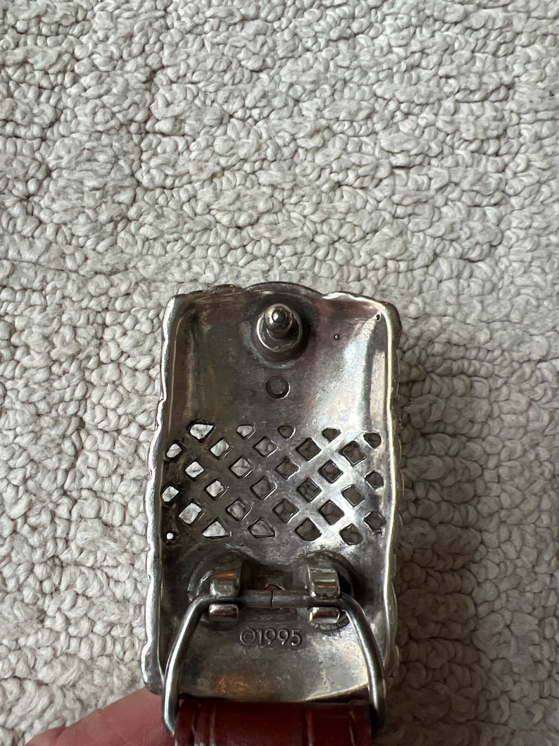 May include: Silver rectangular belt buckle with a decorative lattice pattern and a central stud. The buckle is attached to a brown leather belt. The year "1995" is stamped on the buckle.