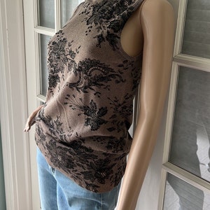 Op de afbeelding: Mouwloze coltrui met een bruin en zwart bloemenpatroon. De top is gemaakt van een gebreide stof en heeft een hoge halslijn. De mannequin draagt ook een blauwe spijkerbroek.