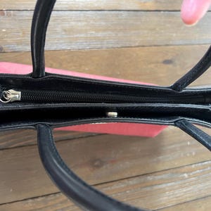 May include: A small, open handbag with a black exterior and black handles. The interior is a light red color. The bag has a zipper closure and a snap closure. The handles are curved and attached to the top of the bag.