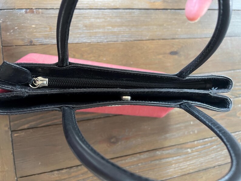 May include: A small, open handbag with a black exterior and black handles. The interior is a light red color. The bag has a zipper closure and a snap closure. The handles are curved and attached to the top of the bag.