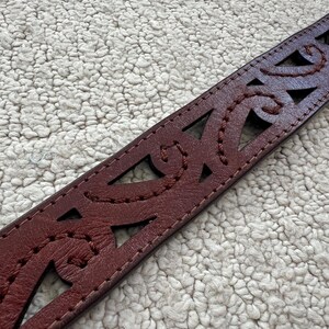 Peut inclure: Gros plan sur une ceinture en cuir bordeaux avec des motifs d&eacute;coup&eacute;s complexes et des coutures d&eacute;coratives. La ceinture pr&eacute;sente des formes g&eacute;om&eacute;triques et des motifs courbes, mettant en valeur un savoir-faire d&eacute;taill&eacute;. C'est un accessoire &eacute;l&eacute;gant.