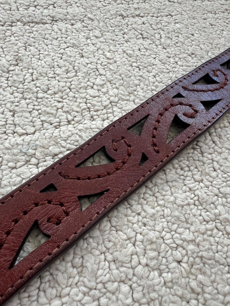 Peut inclure: Gros plan sur une ceinture en cuir bordeaux avec des motifs d&eacute;coup&eacute;s complexes et des coutures d&eacute;coratives. La ceinture pr&eacute;sente des formes g&eacute;om&eacute;triques et des motifs courbes, mettant en valeur un savoir-faire d&eacute;taill&eacute;. C'est un accessoire &eacute;l&eacute;gant.