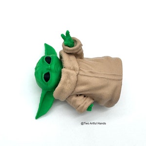 May include: A green figurine of a character with large ears and black eyes, wearing a beige robe. The figurine's hand is raised in a gesture. The text "@Two Artful Hands" is visible.
