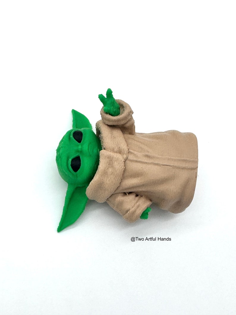 May include: A green figurine of a character with large ears and black eyes, wearing a beige robe. The figurine's hand is raised in a gesture. The text "@Two Artful Hands" is visible.