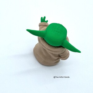 May include: A green and beige figurine of a character with large ears and a small hand raised. The figurine is wearing a beige robe. The text "@Two Artful Hands" is in the bottom right corner.