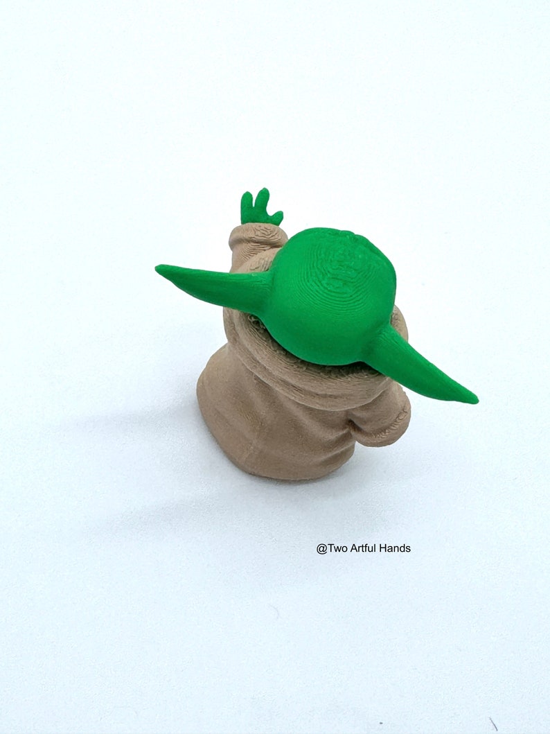 May include: A green and beige figurine of a character with large ears and a small hand raised. The figurine is wearing a beige robe. The text "@Two Artful Hands" is in the bottom right corner.