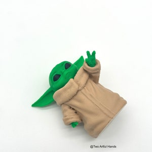 May include: A green figurine of a character with large ears and eyes, wearing a tan robe. The figurine is making a peace sign with its hand. The background is white. The text "@Two Artful Hands" is in the bottom right corner.