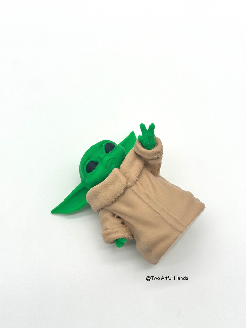 May include: A green figurine of a character with large ears and eyes, wearing a tan robe. The figurine is making a peace sign with its hand. The background is white. The text "@Two Artful Hands" is in the bottom right corner.
