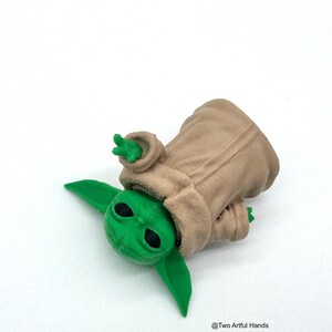 May include: A green figurine of a character from a popular science fiction franchise, wearing a tan robe. The figure has large, black eyes and pointed ears. The figurine is lying on a white surface.