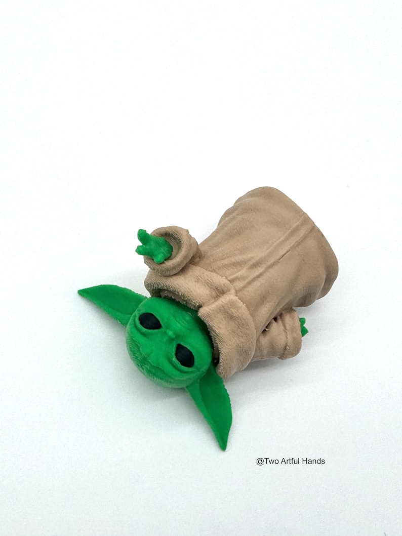 May include: A green figurine of a character from a popular science fiction franchise, wearing a tan robe. The figure has large, black eyes and pointed ears. The figurine is lying on a white surface.