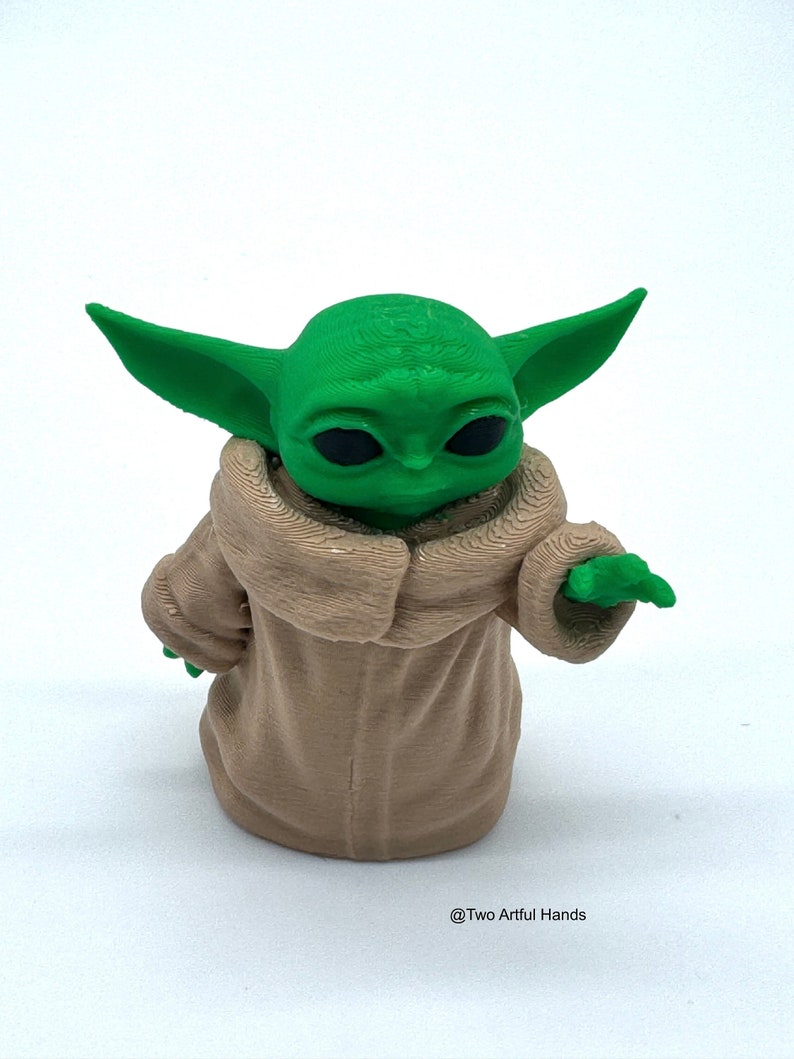 May include: A 3D-printed figurine of a green-skinned character with large ears and black eyes, wearing a tan robe. The figurine is posed with one arm extended. The text "@Two Artful Hands" is at the bottom.