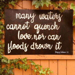 Puede incluir: Un letrero de madera marrón oscuro con letras blancas que dice "many waters cannot quench love, nor can floods drown it" y "Song of Solomon 8.7". El letrero está colgado en una valla de madera con hiedra verde creciendo alrededor.
