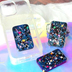 May include: A clear plastic phone case and three rectangular resin grips featuring a dark blue glitter base embedded with multi-colored star-shaped sequins and metallic flakes. The grips have a smooth, glossy finish and a deep, starry appearance.