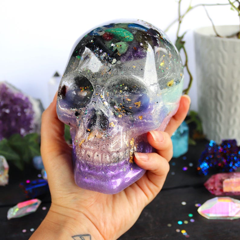 May include: A clear resin skull filled with colorful crystals and glitter. The skull is a light purple color with a silver glitter finish.