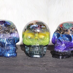 May include: Three crystal skull sculptures with colorful layers of resin and embedded crystals. Each skull has a different color scheme, including blue, green, and purple.