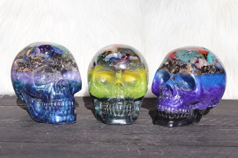 May include: Three crystal skull sculptures with colorful layers of resin and embedded crystals. Each skull has a different color scheme, including blue, green, and purple.