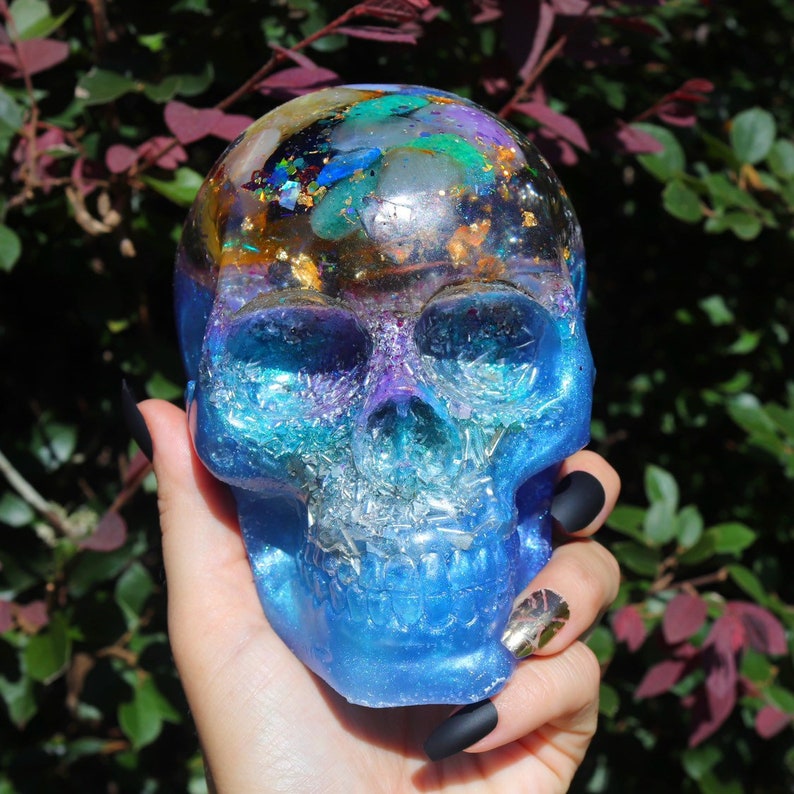 May include: A blue, purple, and gold resin skull with glitter and crystals embedded inside. The skull is sitting in a hand with black nail polish.