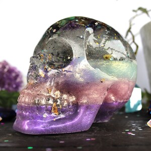May include: A resin skull sculpture with a multi-colored layered design. The skull is decorated with glitter and gold flakes. The colors are purple, pink, blue, and green.