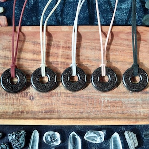 May include: Five black gemstone pendant necklaces with different colored cords: burgundy, white, light pink, and black. The pendants are circular with a hole in the center. The necklaces are displayed on a wooden tray with crystals and gemstones in the background.