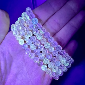 May include: A stack of clear quartz crystal bracelets with a sparkly, iridescent finish. The bracelets are made of round beads and are shown under a UV light, which causes them to glow.