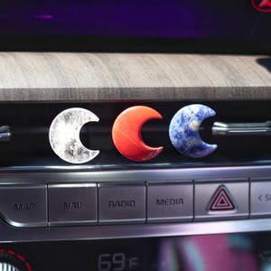 May include: Three crescent moon-shaped car air fresheners. The left one is white with a gray marbled pattern, the middle one is red, and the right one is blue with a white marbled pattern.