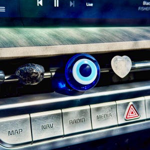 Op de afbeelding: Een auto-interieur met drie decoratieve luchtverfrissers. Een donkergrijze, getextureerde steen, een blauw en wit boze oog en een helder hartvormig kristal worden op het dashboard weergegeven.