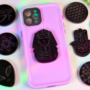 May include: A light pink phone case with a black Tutankhamun phone grip. Surrounding the phone are various black, circular, and hand-shaped decorative items with intricate designs. The items are likely made of resin or a similar material.