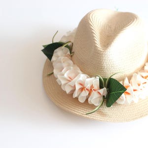 以下が含まれることがあります： 白い花のレイが付いた麦わら帽子。レイはオレンジ色の芯と緑の葉を持つ白い花でできています。