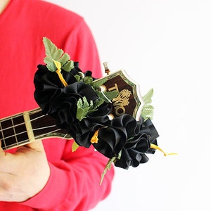 May include: A ukulele headstock adorned with a decorative accessory featuring ruffled black fabric, green artificial fern leaves, and thin yellow ribbons. The wooden headstock displays the gold text TMO with a palm tree graphic above the letters.