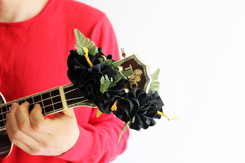 May include: A ukulele headstock adorned with a decorative accessory featuring ruffled black fabric, green artificial fern leaves, and thin yellow ribbons. The wooden headstock displays the gold text TMO with a palm tree graphic above the letters.