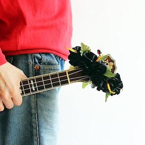 May include: A wooden ukulele with a dark fretboard and silver frets, featuring a decorative cluster of black fabric flowers and green leaves attached to the headstock. The instrument has a natural wood finish and is held horizontally.