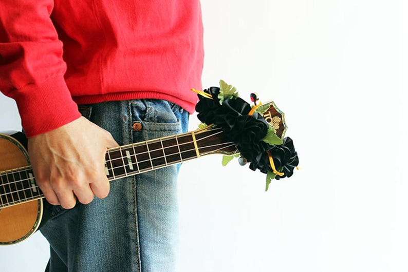 May include: A wooden ukulele with a dark fretboard and silver frets, featuring a decorative cluster of black fabric flowers and green leaves attached to the headstock. The instrument has a natural wood finish and is held horizontally.