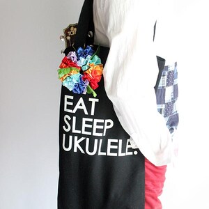 Może przedstawiać: Czarna torba typu tote z napisem "Eat Sleep Ukulele" w kolorze białym. Do torby przymocowany jest kwiat w kolorach tęczy.