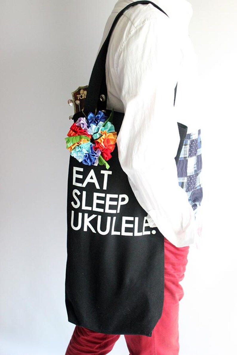 Może przedstawiać: Czarna torba typu tote z napisem "Eat Sleep Ukulele" w kolorze białym. Do torby przymocowany jest kwiat w kolorach tęczy.