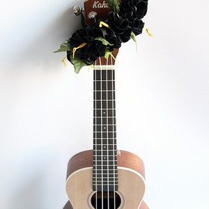 May include: A light brown wooden ukulele with a dark fretboard and four strings. The headstock is decorated with a cluster of black fabric flowers, green artificial leaves, and thin yellow ribbons. The brand name Kahu is visible on the headstock.