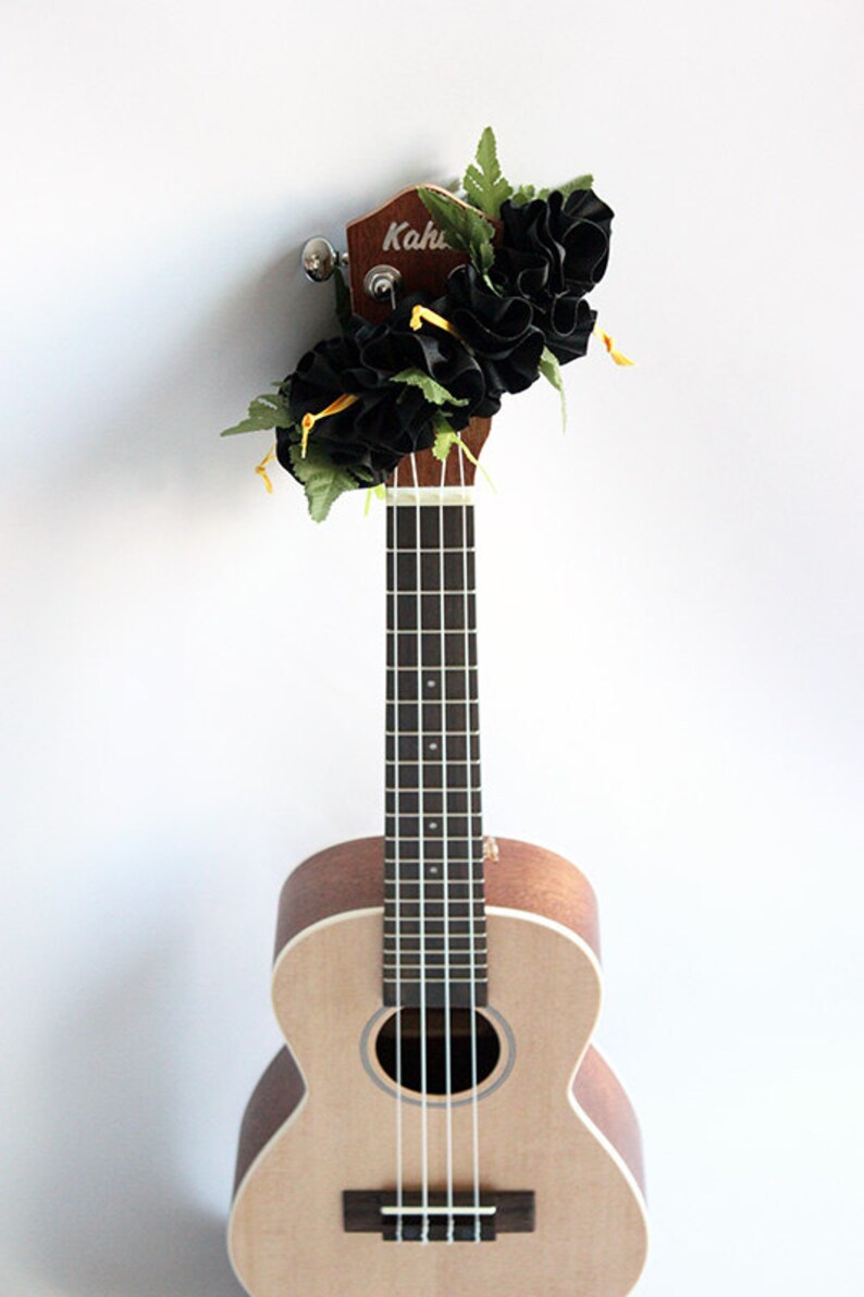 May include: A light brown wooden ukulele with a dark fretboard and four strings. The headstock is decorated with a cluster of black fabric flowers, green artificial leaves, and thin yellow ribbons. The brand name Kahu is visible on the headstock.