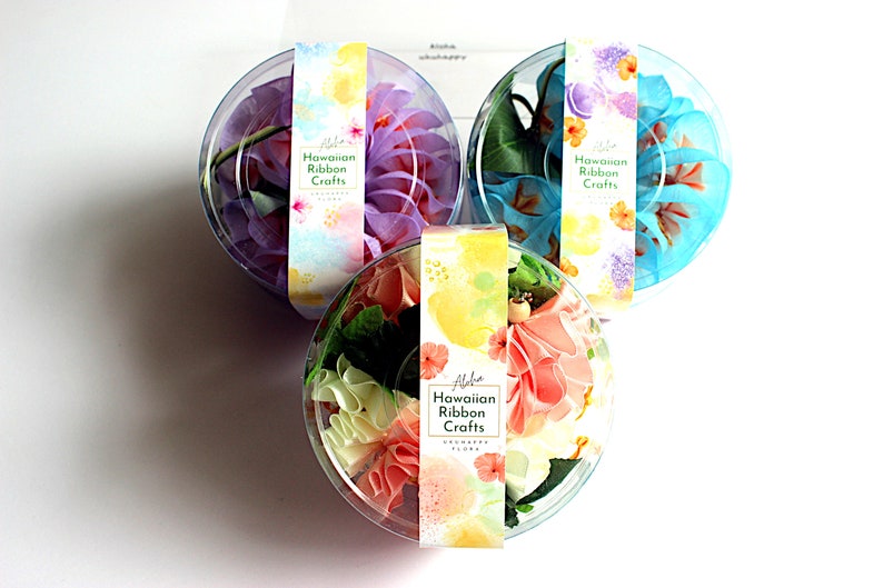 May include: Three circular clear plastic containers holding fabric ribbon flowers in purple, blue, and peach. Each container features a paper label with the text Hawaiian Ribbon Crafts and Uohappy Flora, decorated with colorful watercolor floral patterns.