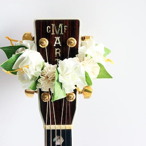 以下が含まれることがあります： C, M, A, Rの文字が象嵌されたアコースティックギターのヘッドストック。白い紙の花と緑の葉のフローラルガーランドで飾られています。金色のチューニングペグと弦が見えます。