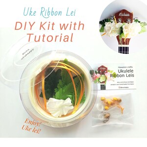 May include: A DIY kit for making a ukulele ribbon lei. The kit includes white ribbon, green leaves, a white flower, and yellow beads. The kit comes in a clear plastic container with a lid. The image also shows a ukulele with a lei made from the same materials.