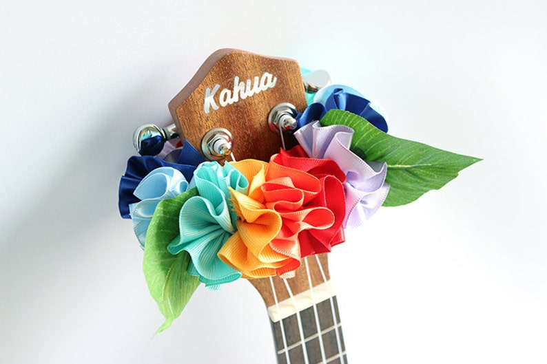 Może przedstawiać: Ukulele z wiankiem z kwiat&oacute;w tęczy wykonanym z wstążki wok&oacute;ł gł&oacute;wki. Gł&oacute;wka jest drewniana i ma napis "Kahua".