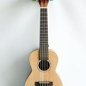 Może przedstawiać: Ukelele z jasnego drewna z tęczowym lei na gł&oacute;wce. Ukelele ma czarną podstrunnicę i białe struny. Na gł&oacute;wce widnieje napis "Kahua".