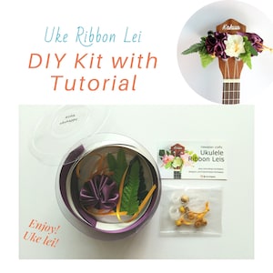 May include: A DIY kit for making a ukulele ribbon lei. The kit includes purple ribbon, yellow ribbon, green fern leaves, a purple flower, and a small bag of yellow beads. The kit is packaged in a clear plastic container with a lid. The image also shows a ukulele with a purple and white ribbon lei attached to the headstock. The text on the card reads "Hawaiian crafts Ukulele Ribbon Leis".