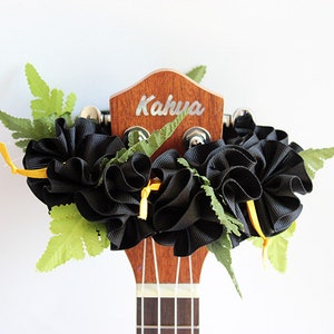 May include: A wooden ukulele headstock with the brand name Kahua printed in white. The headstock is adorned with a decorative arrangement of black ruffled ribbon, small yellow ribbon ties, and artificial green fern leaves attached to the tuning pegs.