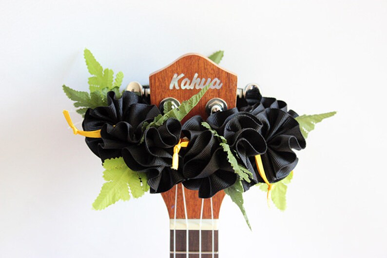 May include: A wooden ukulele headstock with the brand name Kahua printed in white. The headstock is adorned with a decorative arrangement of black ruffled ribbon, small yellow ribbon ties, and artificial green fern leaves attached to the tuning pegs.