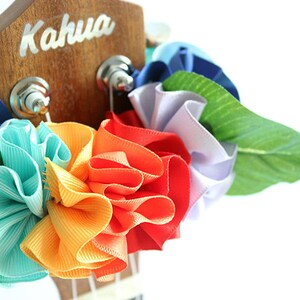 Może przedstawiać: Ukulele ozdobione tęczowym lei z kolorowych kwiat&oacute;w z wstążek. Głowica ukulele jest drewniana i ma napis "Kahua".