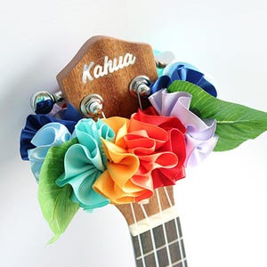 Może przedstawiać: Ukulele z tęczowym lei wykonanym z kolorowych kwiat&oacute;w z tkaniny i zielonych liści. Głowica ukulele jest oznaczona napisem "Kahua".
