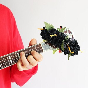 May include: A wooden ukulele headstock adorned with a decorative arrangement of black fabric flowers and green artificial fern leaves. The instrument features silver tuning pegs and a dark fretboard with white position markers on the neck.