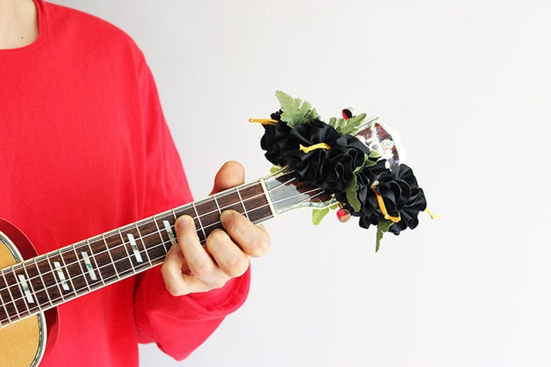 May include: A wooden ukulele headstock adorned with a decorative arrangement of black fabric flowers and green artificial fern leaves. The instrument features silver tuning pegs and a dark fretboard with white position markers on the neck.